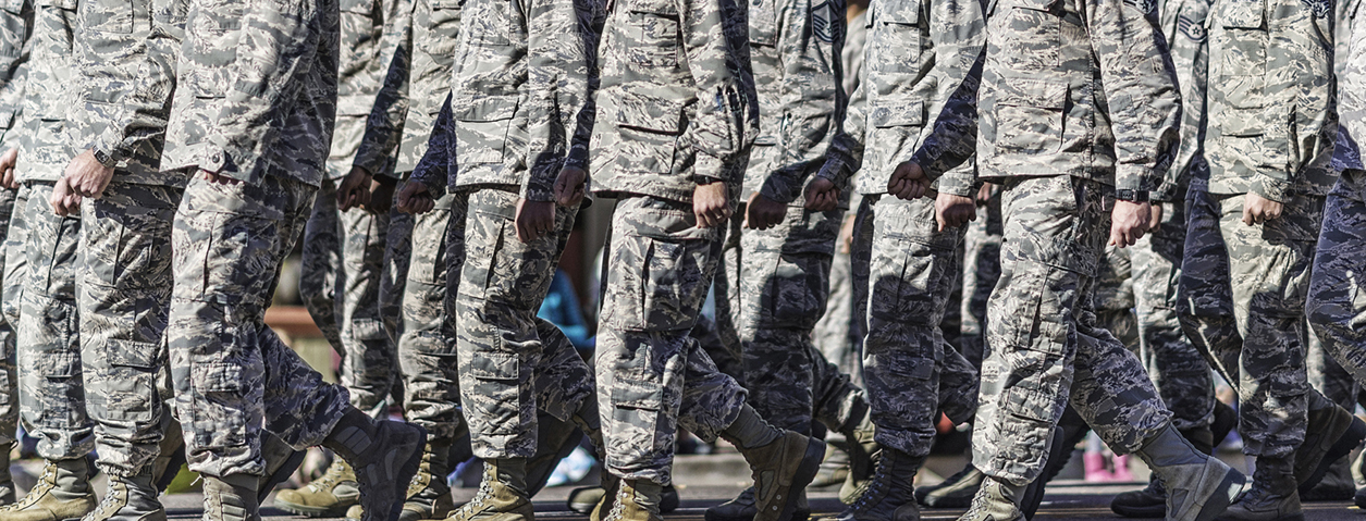A close-up view of a large group of U.S. military personnel marching in formation, showing their torsos and legs from the waist down. All are wearing matching camouflage uniforms and tan combat boots, with arms swinging in unison. The image is cropped to emphasize the uniformity and synchronized movement of the formation.