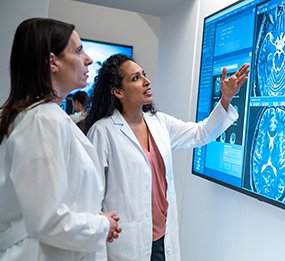 Two female doctors review digital brain scans on a large wall monitor.]