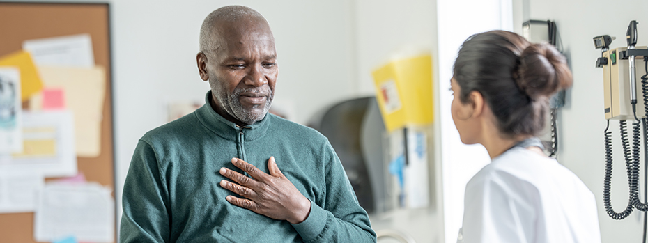 An older man sits on a medical exam table with his hand on his heart as he talks to a young female doctor.