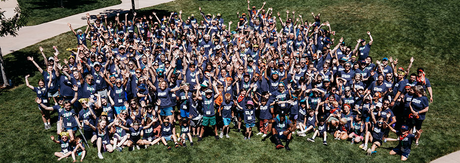A large group of more than 100 people in matching t-shirts pose on the grass and wave.