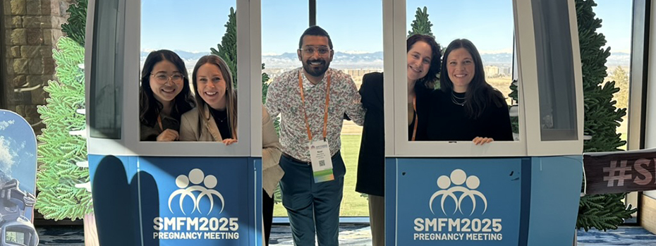 A man standing with two woman on his left side and two other woman on his right side, all smiling, in a lift car that says "SMFM2025 Pregnancy Meeting" on the front.