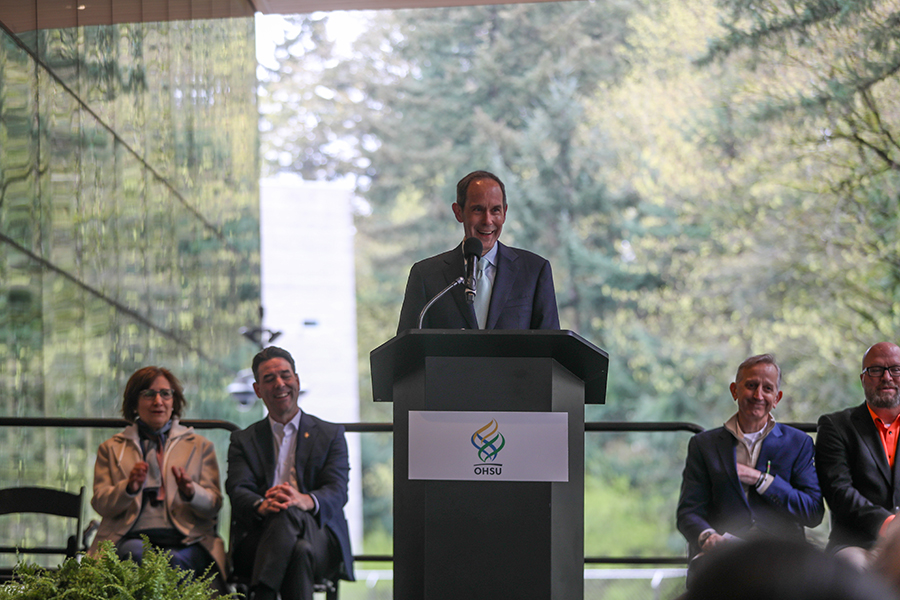 Dr. Brian Druker standing at a podium with the OHSU logo on the front with four people seated in a row behind him.