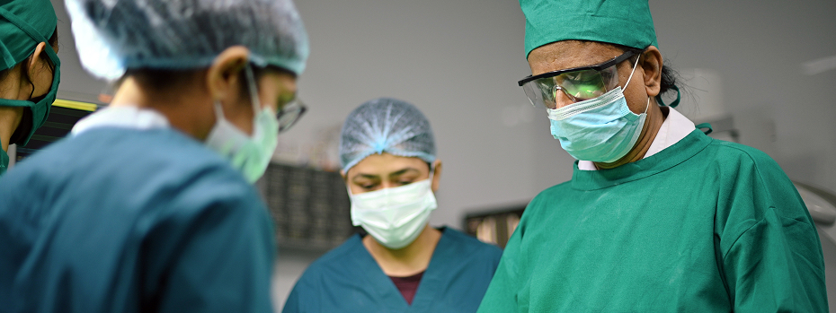 A senior male surgeon performs surgery in an operating room with two female surgeons assisting.