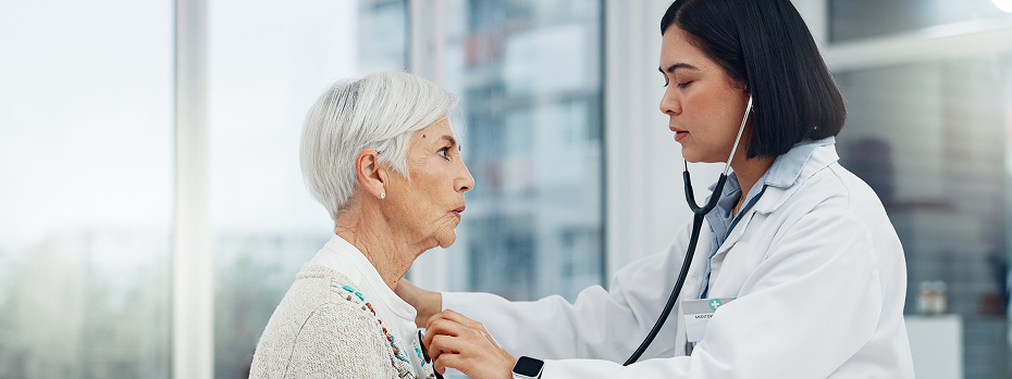 Female doctor uses a stethoscope to check an older woman’s heart in a medical exam room.