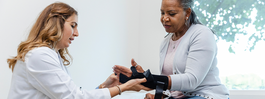 A female doctor in a medical exam room helps an older woman adjust her orthopedic wrist brace.
