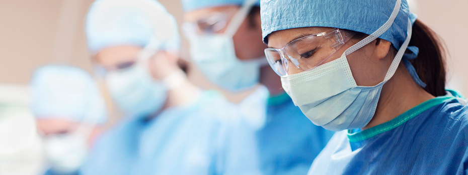A female surgeon wearing a mask looks down at a patient in a hospital operating room with a team of colleagues.