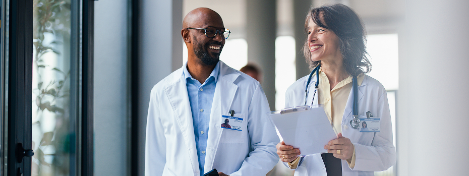 A male and a female doctor smile and talk as they walk along a hallway lined with windows.
