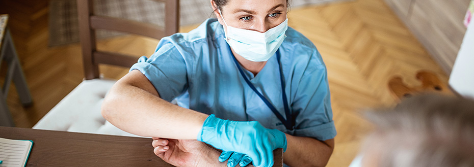 A nurse wearing a mask takes a blood sample from an older patient at her home.