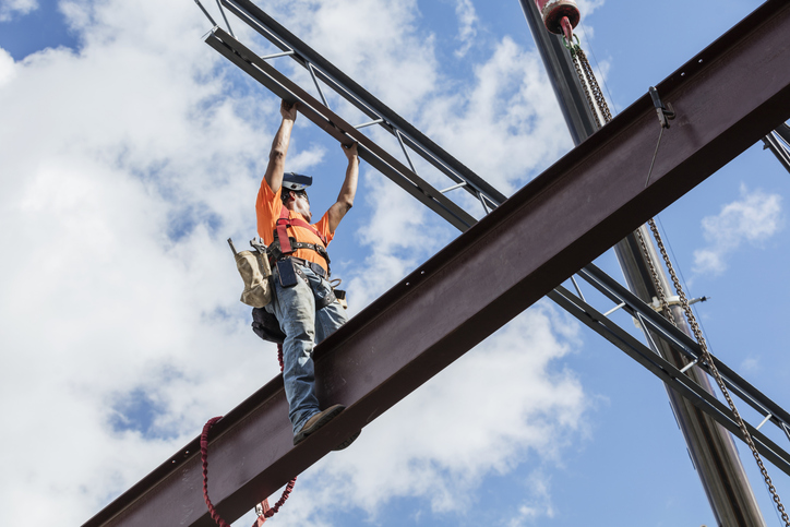 Claude responded: A construction ironworker is standing on a steel beam high above the ground, wearing an orange safety vest, hard hat, fall protection harness, and tool belt.A construction ironworker is standing on a steel beam high above the ground, wearing an orange safety vest, hard hat, fall protection harness, and tool belt. They're reaching upward to guide or connect another steel beam, with a blue cloudy sky in the background. A chain (likely from a crane) is visible on the right side.