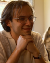 A man smiling with his elbows on a table and his hands together under his chin.