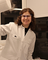 A woman wearing a white lab coat and protective goggles smiles while holding up a sample that is emitting smoke in a research lab.