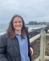 A woman leaning against a fence smiling in front of a beach on a cloudy day.