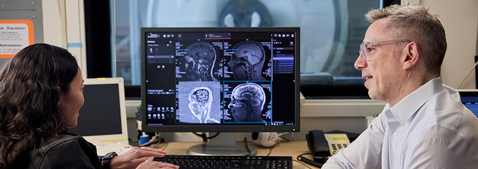 Dr. Yamamoto and Dr. Piantino sit in front of a computer screen showing images of a brain MRI in progress. In the background through a large window, we see the MRI machine with a patient inside.