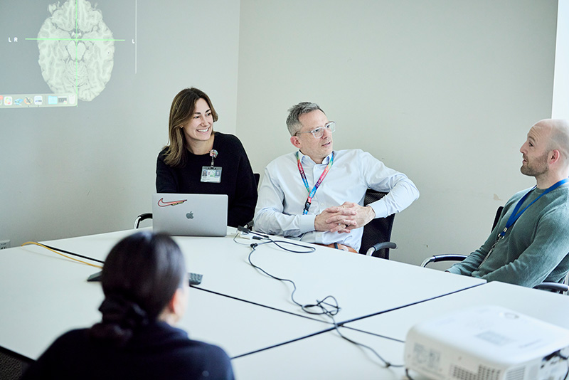 Dr. Piantino and three lab team members sit and talk at a large white table. A projector screen on the wall shows a brain scan image.