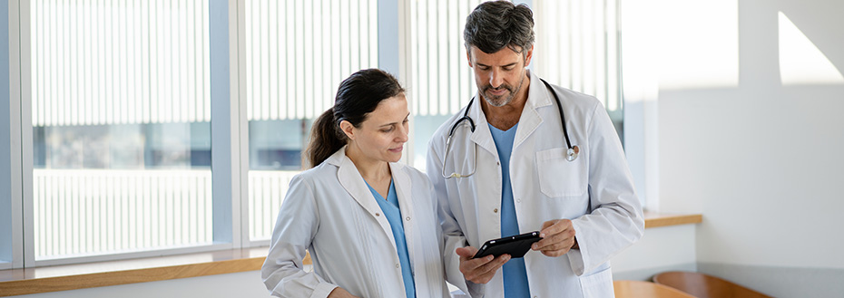 Two doctors in white coats look at a tablet and discuss patient care in a hospital hallway.