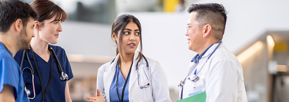 Four medical professionals stand in the hallway of a hospital. They are each dressed professionally in scrubs and white lab coats as a female doctor speaks to her colleagues.
