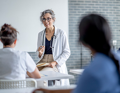 A mature female doctor sits at the front of a classroom as she instructs a small group of students