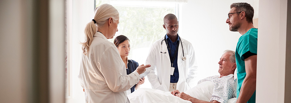 A group of medical professionals stand around an older male patient laying in a hospital bed.
