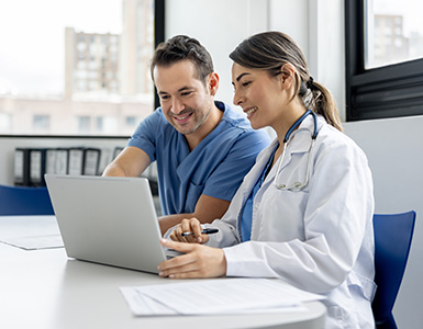 An older male doctor and a younger female doctor talk and work on a laptop in a conference room.