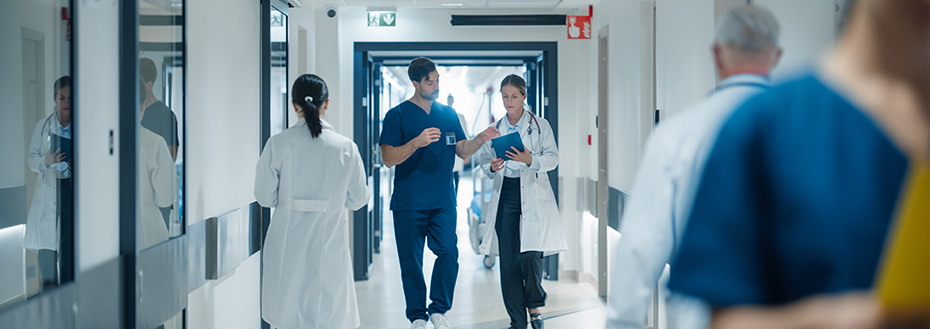 Two doctors walk through a busy hospital hallway going over notes on a clipboard.