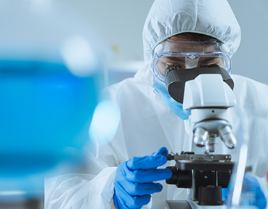 A male researcher in PPE looks into a microscope in a laboratory.
