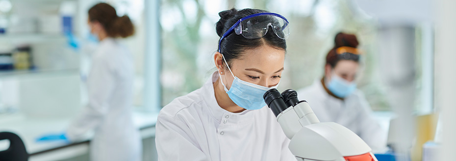 A female researcher wearing protective gear looks through a microscope in a research lab.