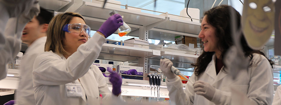 In a lab, a scientist wearing safety glasses holds up a flask containing a sample. Another scientist holds a vial.