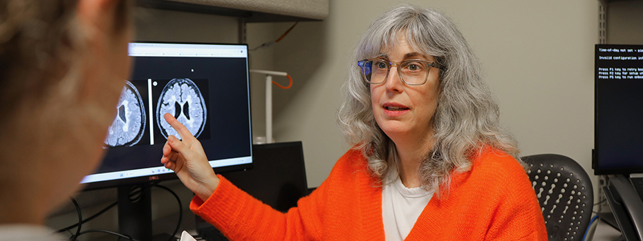 A woman sitting in front of a computer points to the monitor that's displaying a brain scan as she looks at another person sitting next to her.