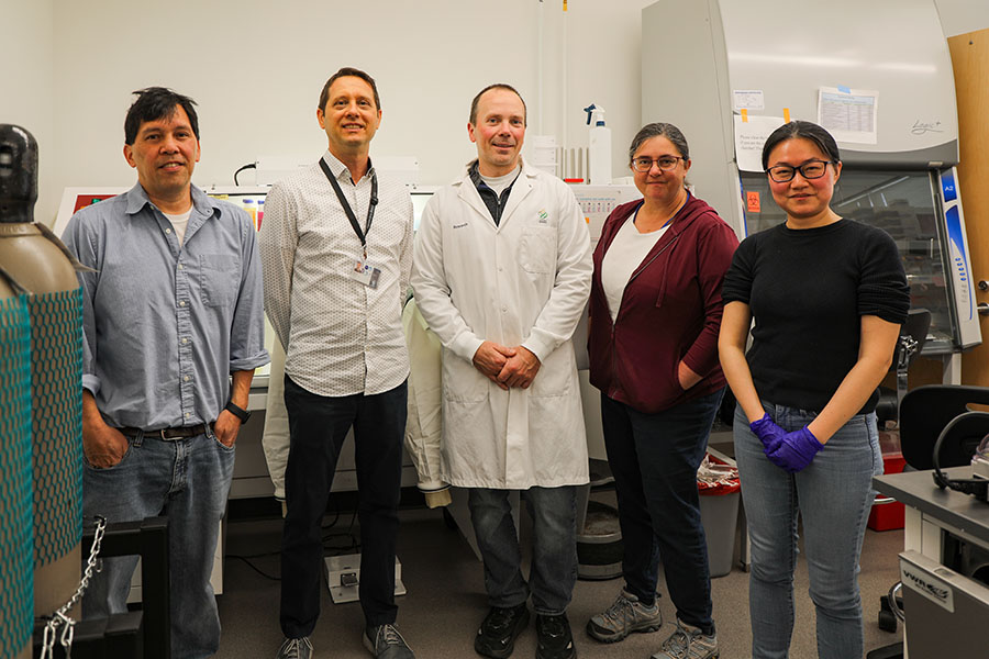 A man wearing a white lab coat stands in a research lab with two men on his right side and two women on his left side.