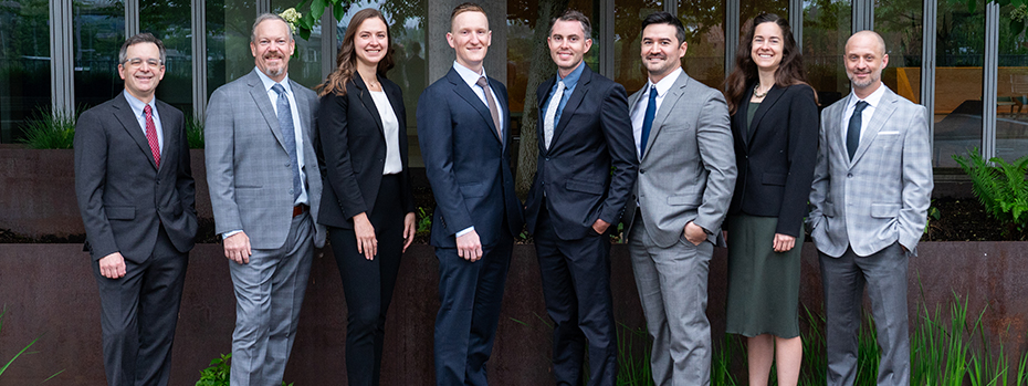 Six men and two women stand next to each other smiling outside in front of a window, all nicely dressed.
