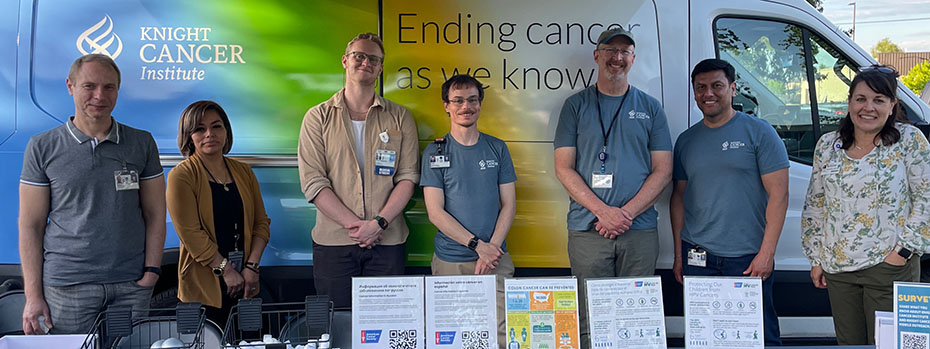 Seven people stand behind a table with health education materials. They are in front of a van labeled “Knight Cancer Institute: Ending cancer as we know it.”