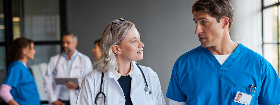 Healthcare professionals in a hospital corridor walk and talk while reviewing information on a tablet, with additional medical staff in the background.