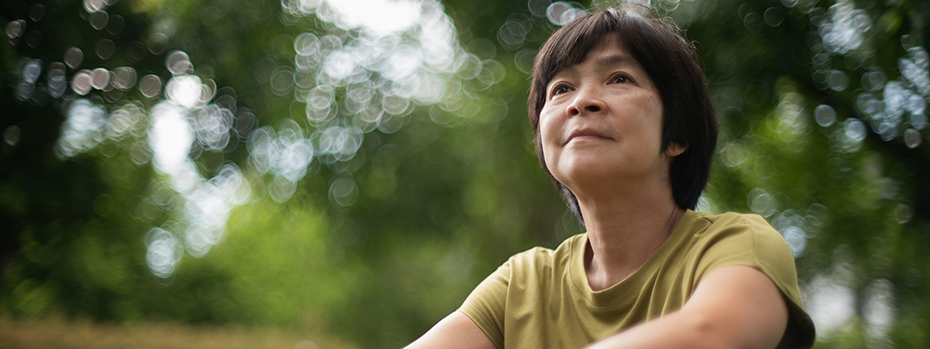 A mature Asian woman sits in a park, looking into the distance.