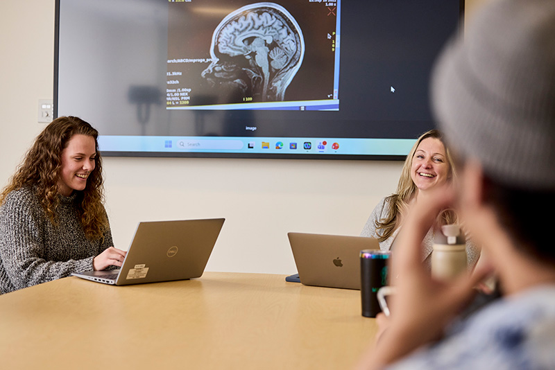 Harkness and Dr. Huber sit at a conference table with laptops in front of them. A screen behind them shows a brain scan image. The back of another team member is blurry in the foreground.