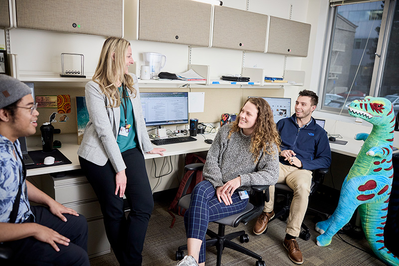 Anthony Hill, Dr. Rebekah Huber, Anya Harkness and Sam Sievertsen sit or stand in front of a row of desks with computer screens. Next to them stands a four-foot-tall inflatable dinosaur.