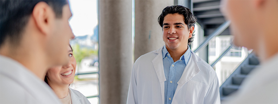 Four PA students in white coats talk together in a hallway.