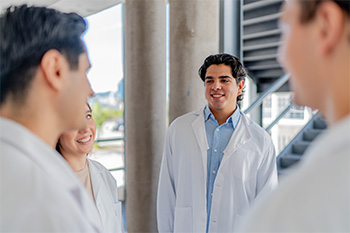 Four PA students in white coats talk together in a hallway. 