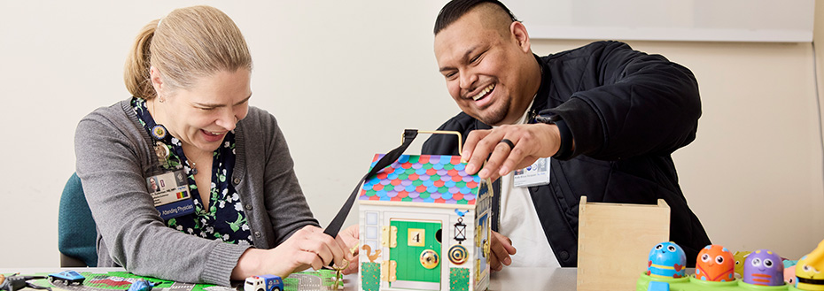 Dr. Zuckerman and Rivas Vazquez sit at a table, laughing and playing with a colorful toy house. There are puzzles, trucks and blocks on the table in the foreground.