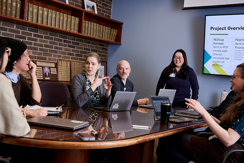 6 team members are visible around a conference table. Dr. Zuckerman speaks with the group. Several team members have laptops on the table and a screen on the wall behind the table shows an overview of the project they’re discussing.