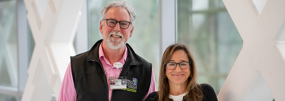 A man wearing an OHSU-branded vest and a women pose smiling in front of windows.