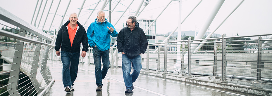 Three senior men in rain jackets walk across a bridge in Portland.