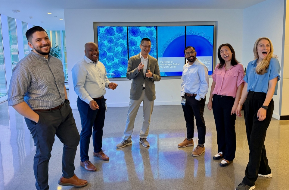 A group of six people stand in a semi-circle smiling at the camera. There is a large screen behind them showing stem cells.