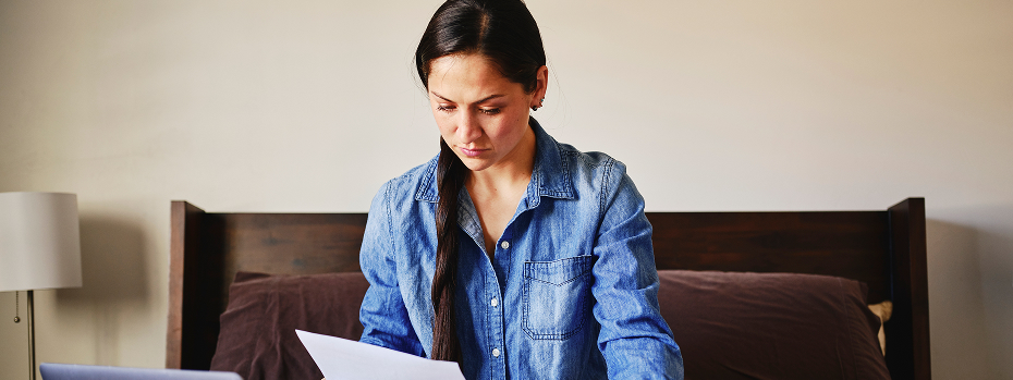 A young woman sits on her bed working with her laptop and medical papers.