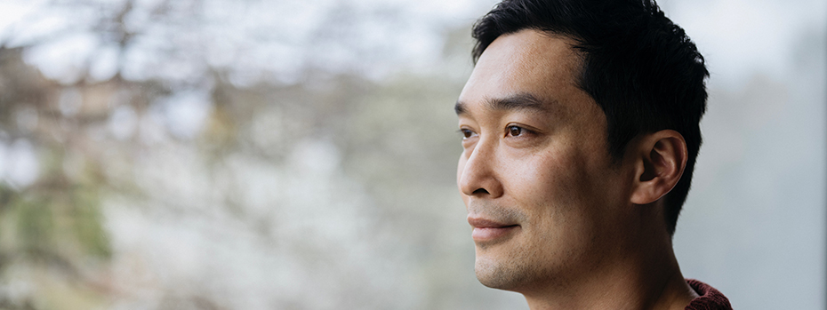 An Asian man stands outside and looks into the distance with a calm expression.