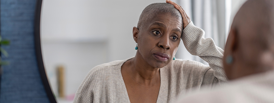 A Black woman with close-cropped hair looks into a mirror with a concerned expression and one hand on her head.