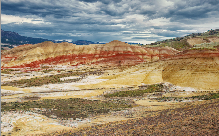 Photograph of a colorful landscape featuring layered hills with distinct bands of red, yellow, and beige sediment. The scene includes a partly cloudy sky and distant mountains, highlighting natural geological formations and erosion patterns.