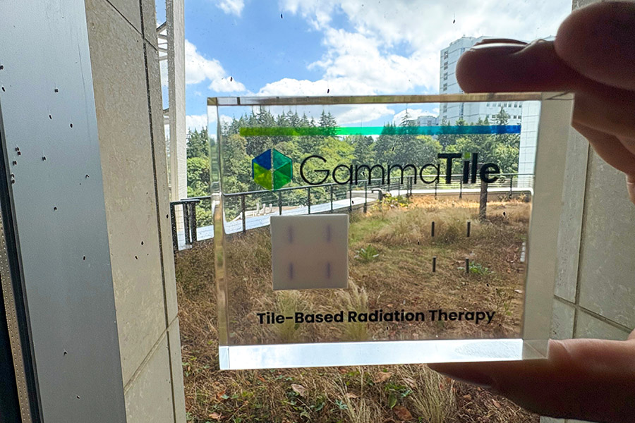 Hands holding up a clear plaque that has the GammaTile logo on it and says "Tile-Based Radiation Therapy," with part of OHSU campus visible through the plaque.