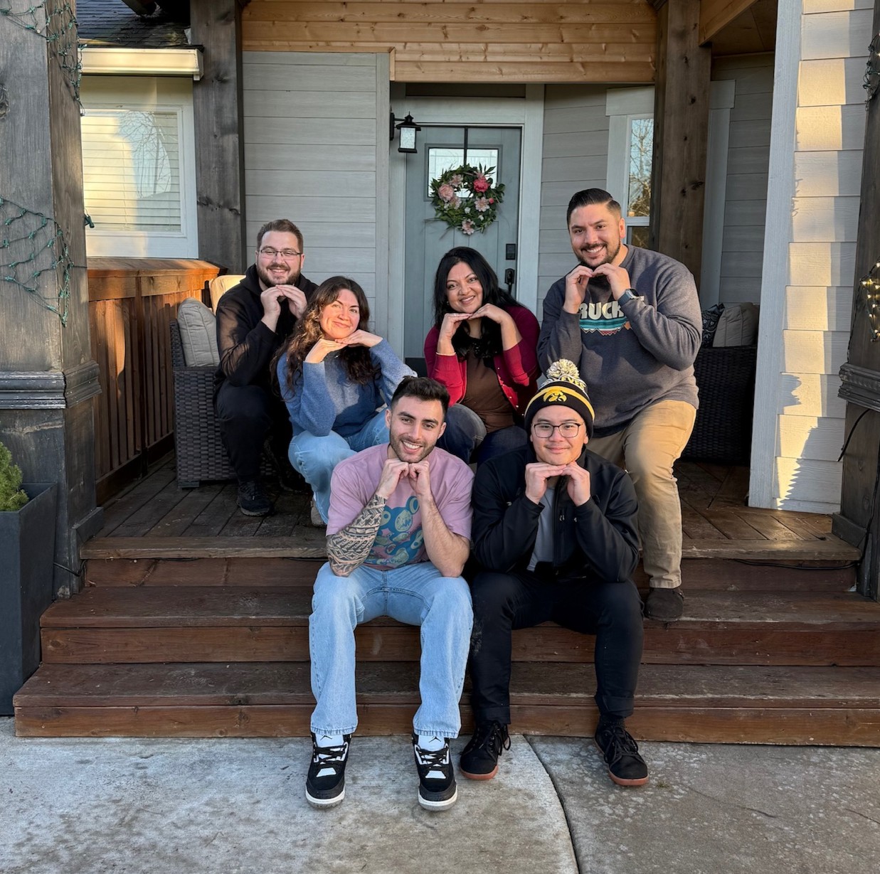 A group of 6 people pose with ands under their chins on the steps of a house.