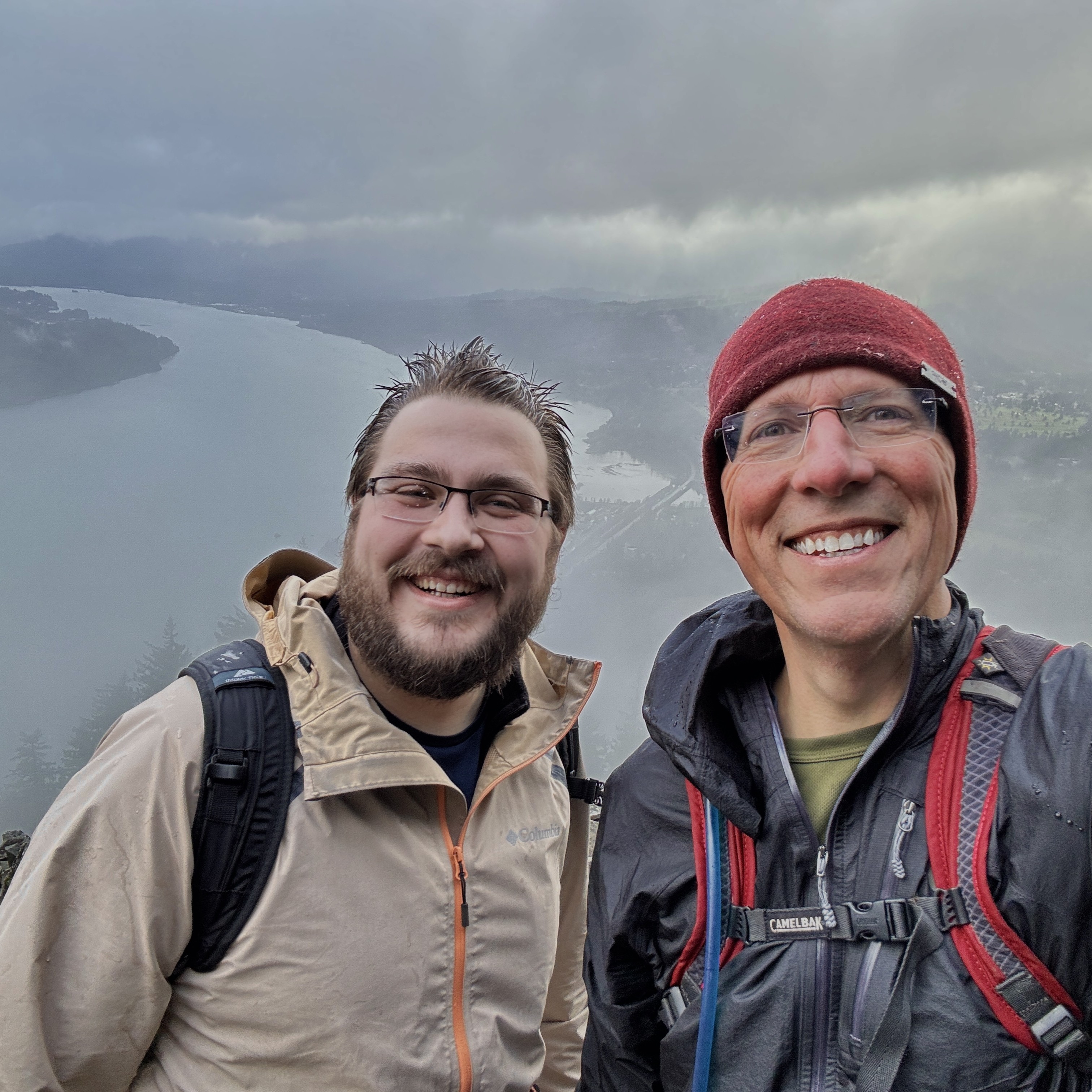 Two people pose for a selfie on a rainy cold day with a sweeping view of mountains and rivers behind them.