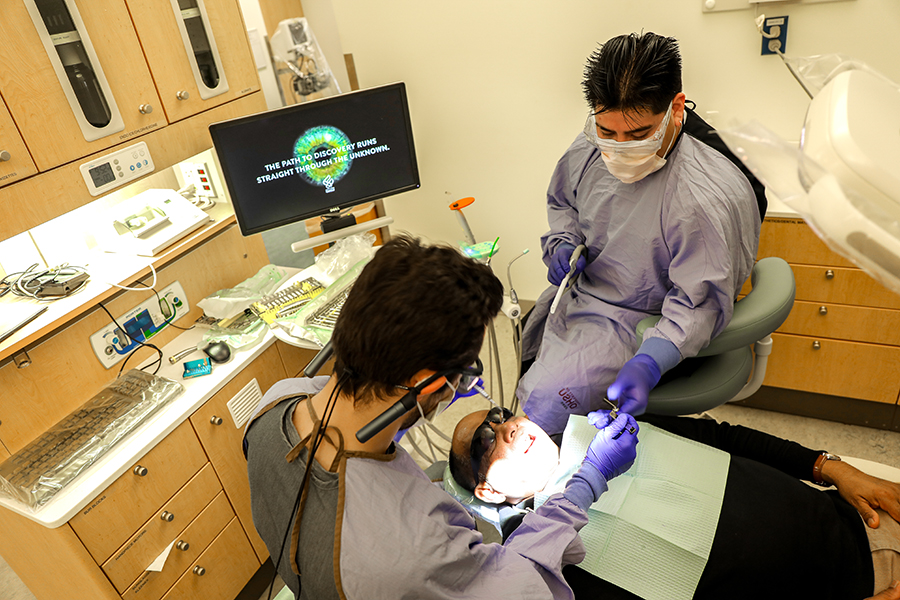 Two people in masks, gloves and gowns sit on either side of a dental patient.